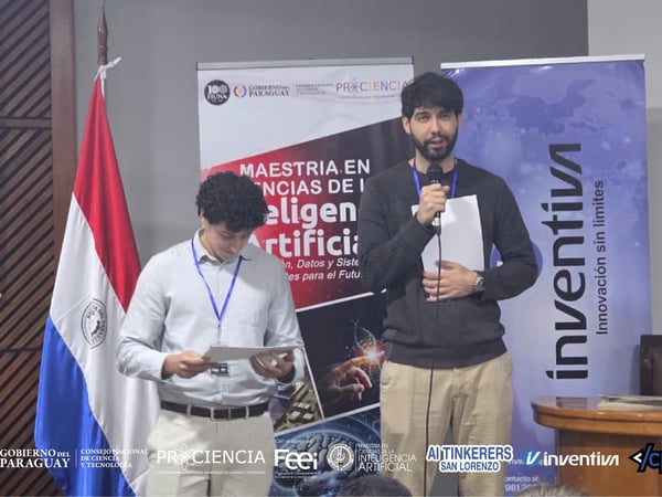 Two men are on a stage during a presentation for an Artificial Intelligence Master's program in Paraguay, standing in front of event banners and the national flag.