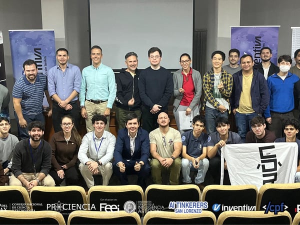 A large group of students and faculty posing for a commemorative group photo in an auditorium, featuring several event banners and sponsor logos.