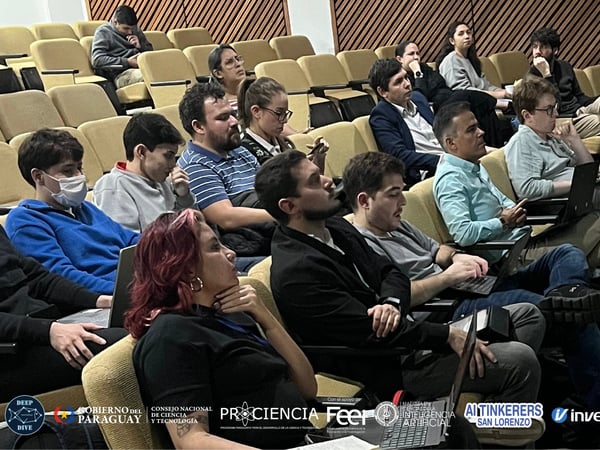 An audience of young adults sits in a tiered auditorium, many with laptops open, focused on a presentation occurring off-camera.