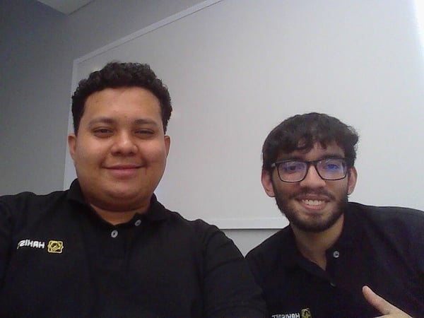 Two men wearing matching black polo shirts are smiling for a photo in front of a white whiteboard in an indoor setting.