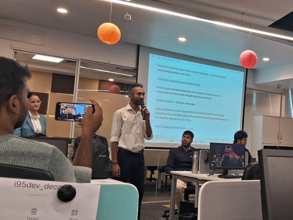 A man stands in a modern office setting, speaking into a microphone during a presentation with a projector screen displaying technical information behind him.