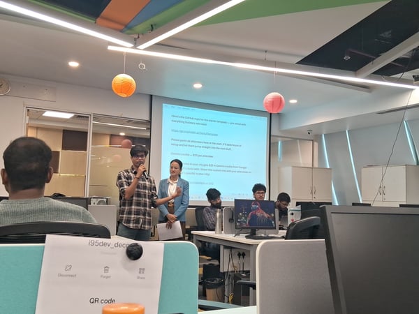 A man stands at the front of a modern office space giving a presentation with a projection screen behind him, while several people listen and work at their desks.