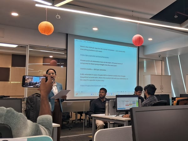 A woman delivers a presentation in an office setting while a person in the foreground records her with a smartphone.