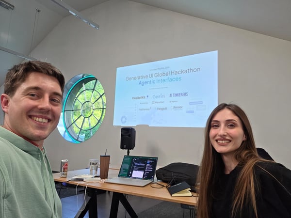 A man and a woman are smiling for a selfie in an indoor setting, with a projection screen in the background announcing a 'Generative UI Global Hackathon'.