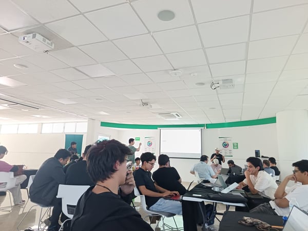 A group of people are gathered in a large room for a presentation or workshop, with many participants sitting at tables using laptops while others stand near a projection screen.