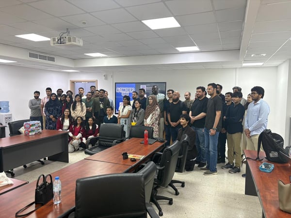 A group photo of event attendees posing together in a modern conference room with wooden tables and a display screen.