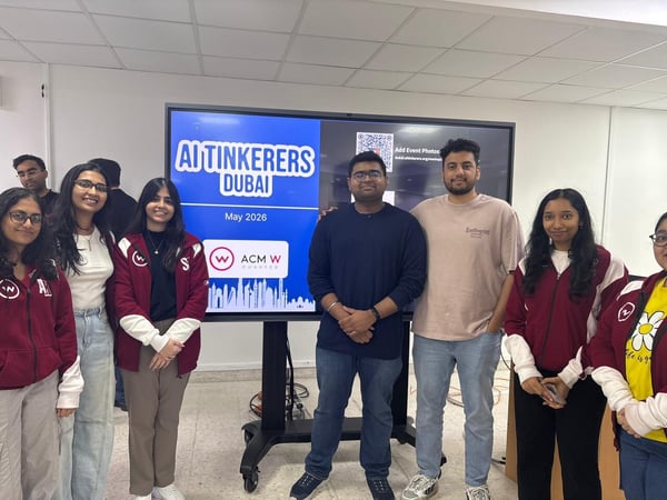 A group of young adults posing together indoors in front of a digital screen displaying event information.