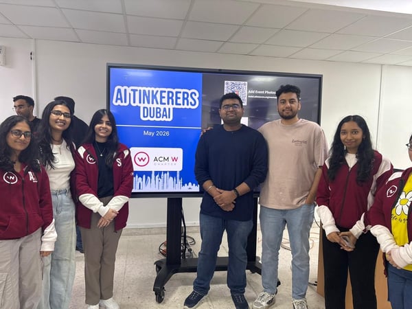 A group of young adults posing together indoors in front of a large digital screen displaying event details.