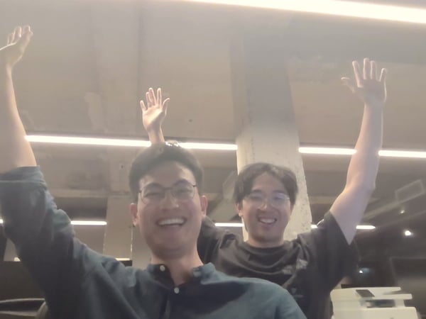 Two young men in an office environment smile brightly while raising their arms above their heads in a celebratory pose. The photograph is heavily overexposed due to bright overhead lighting.